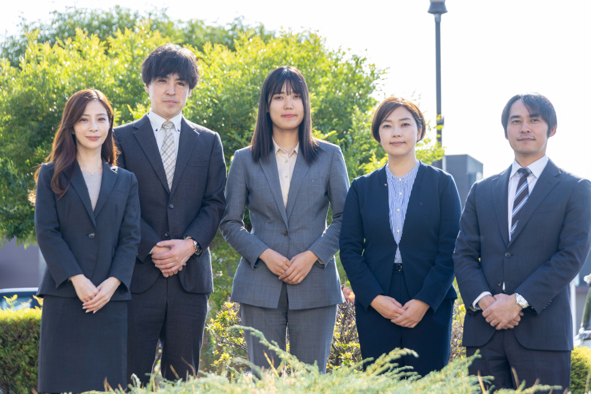 緑豊かな屋外を背景に、スーツを着用した5人の若手アジア系ビジネスパーソン（男性2人、女性3人）が並んで立つ集合写真。全員が真剣な表情で前を見つめており、新入社員、プロジェクトチーム、または企業のプロフェッショナルなイメージを象徴している。