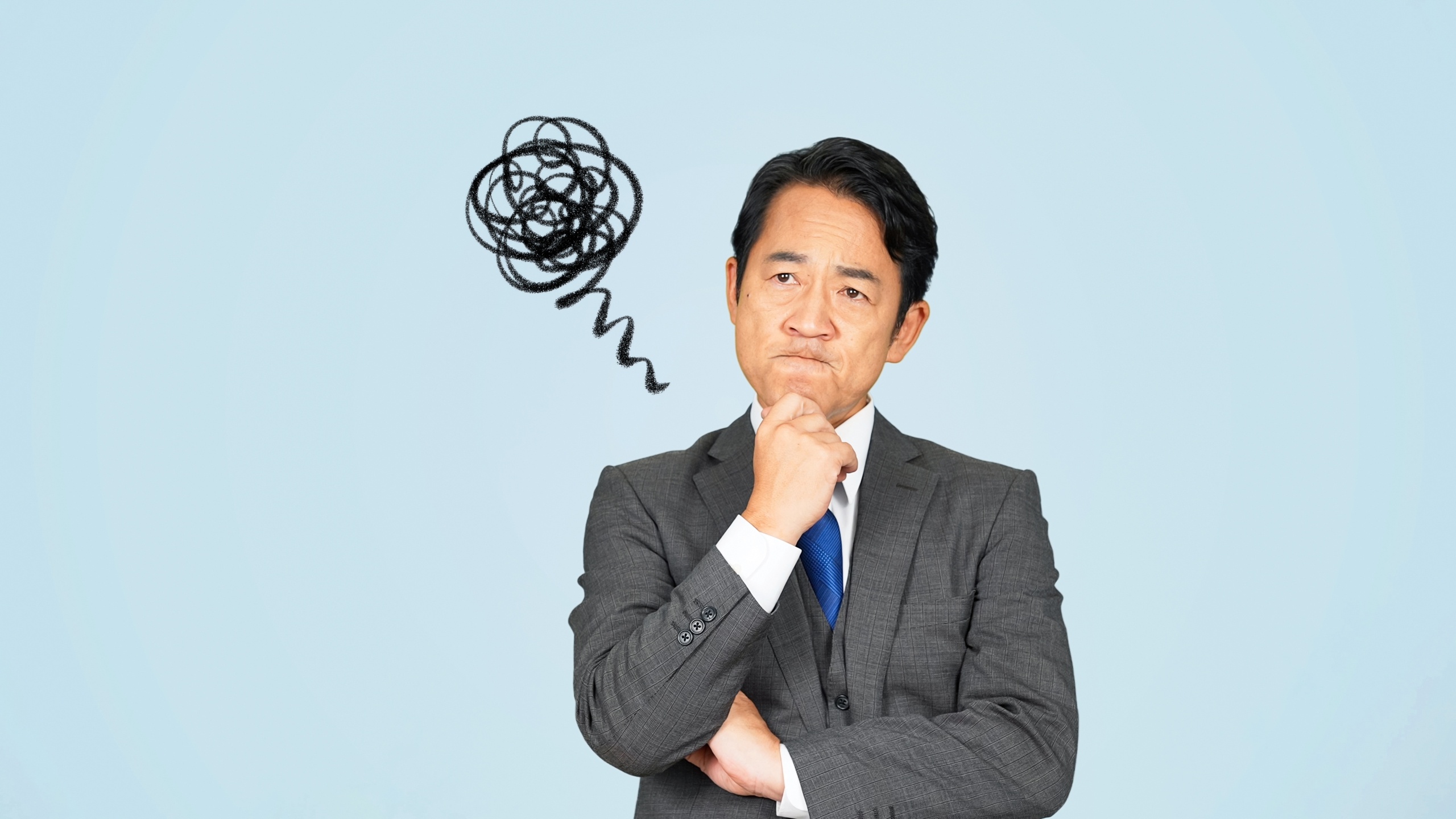 スーツを着た中年のビジネスマンが、顎に手を当てて難しい表情で考え込んでいる。頭の横には、混乱した思考や解決できない悩みを象徴する、黒くもやもやした手書きの線が描かれている。