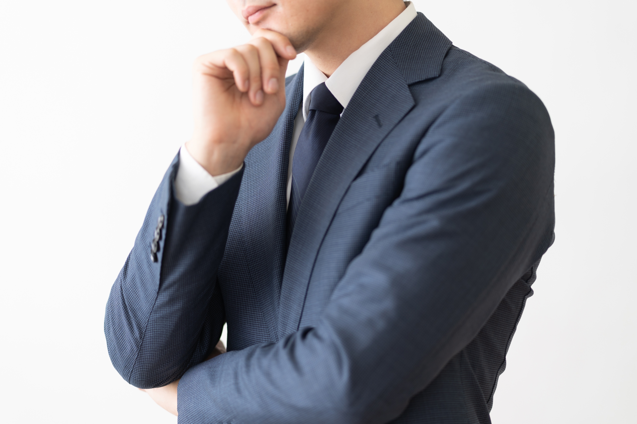 白い背景を前に、ネイビーのスーツを着たビジネスマンが腕を組み、顎に手を当てて思案している。顔の上部は写っておらず、戦略、計画、問題解決といったビジネス上の思考を象徴するクローズアップ写真。
