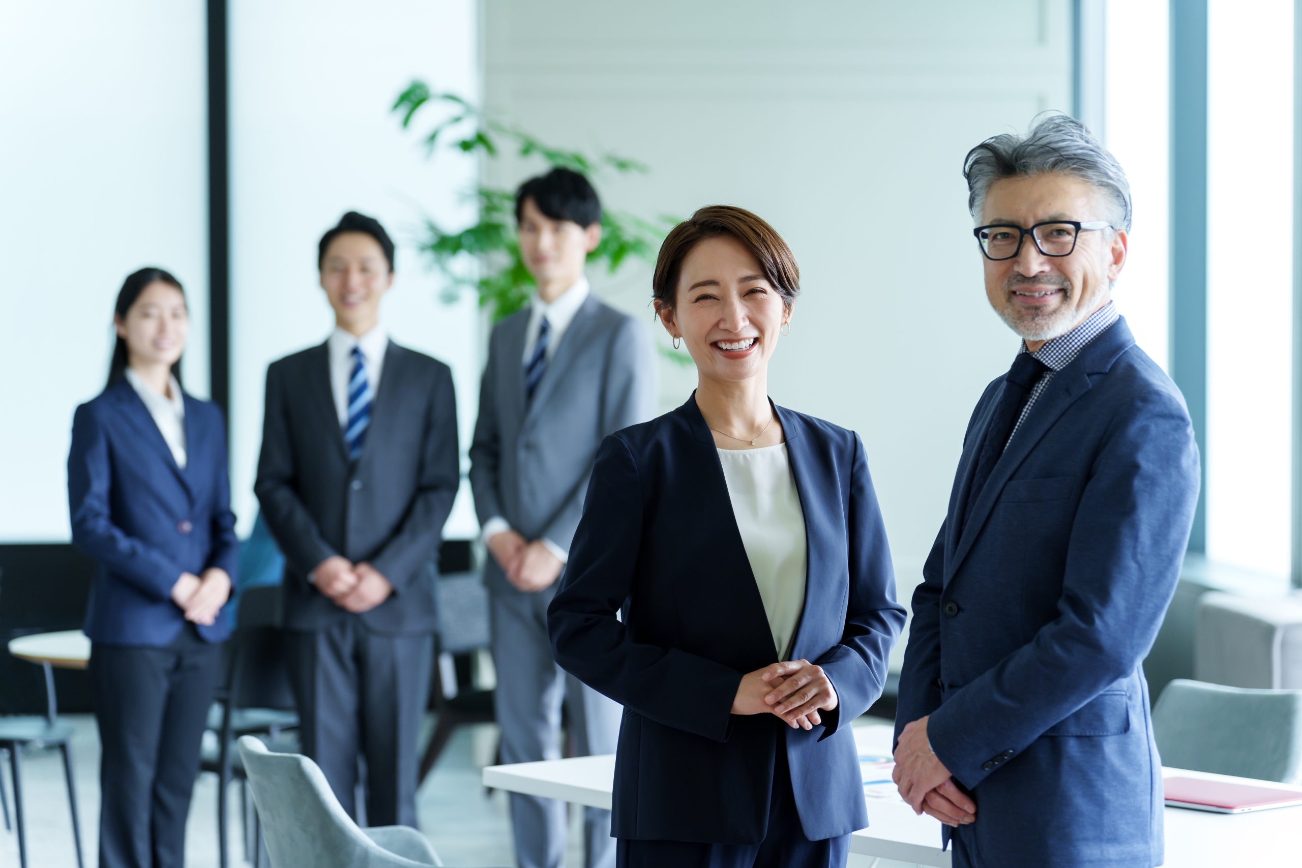 明るい近代的なオフィスで、笑顔でカメラを見つめるアジア系のビジネスチーム5人。最前列にはリーダーシップを感じさせる中年女性と経験豊富な年配男性が立ち、背景には3人の若手社員が並んでいる。チームワークとプロフェッショナリズムを象徴する集合写真。