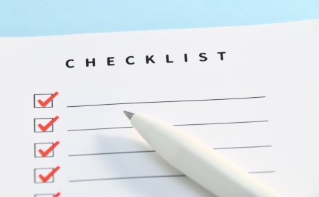 「CHECKLIST」と書かれた白い紙に、複数の項目がリストアップされ、そのうちいくつかのチェックボックスに赤いチェックマークが付けられている。手前には白いペンが置かれており、完了したタスクや計画の実行を示唆している。
