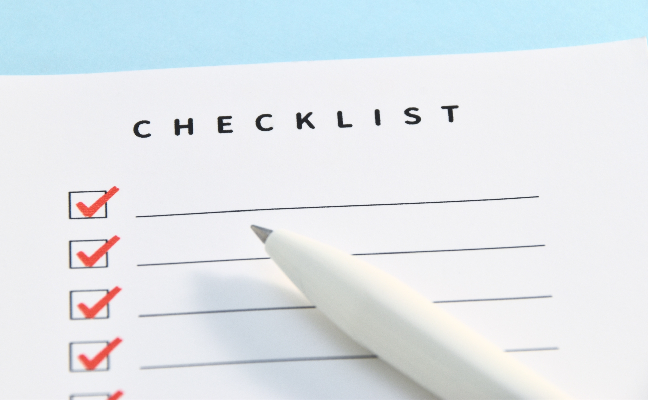 「CHECKLIST」と書かれた白い紙に、複数の項目がリストアップされ、そのうちいくつかのチェックボックスに赤いチェックマークが付けられている。手前には白いペンが置かれており、完了したタスクや計画の実行を示唆している。