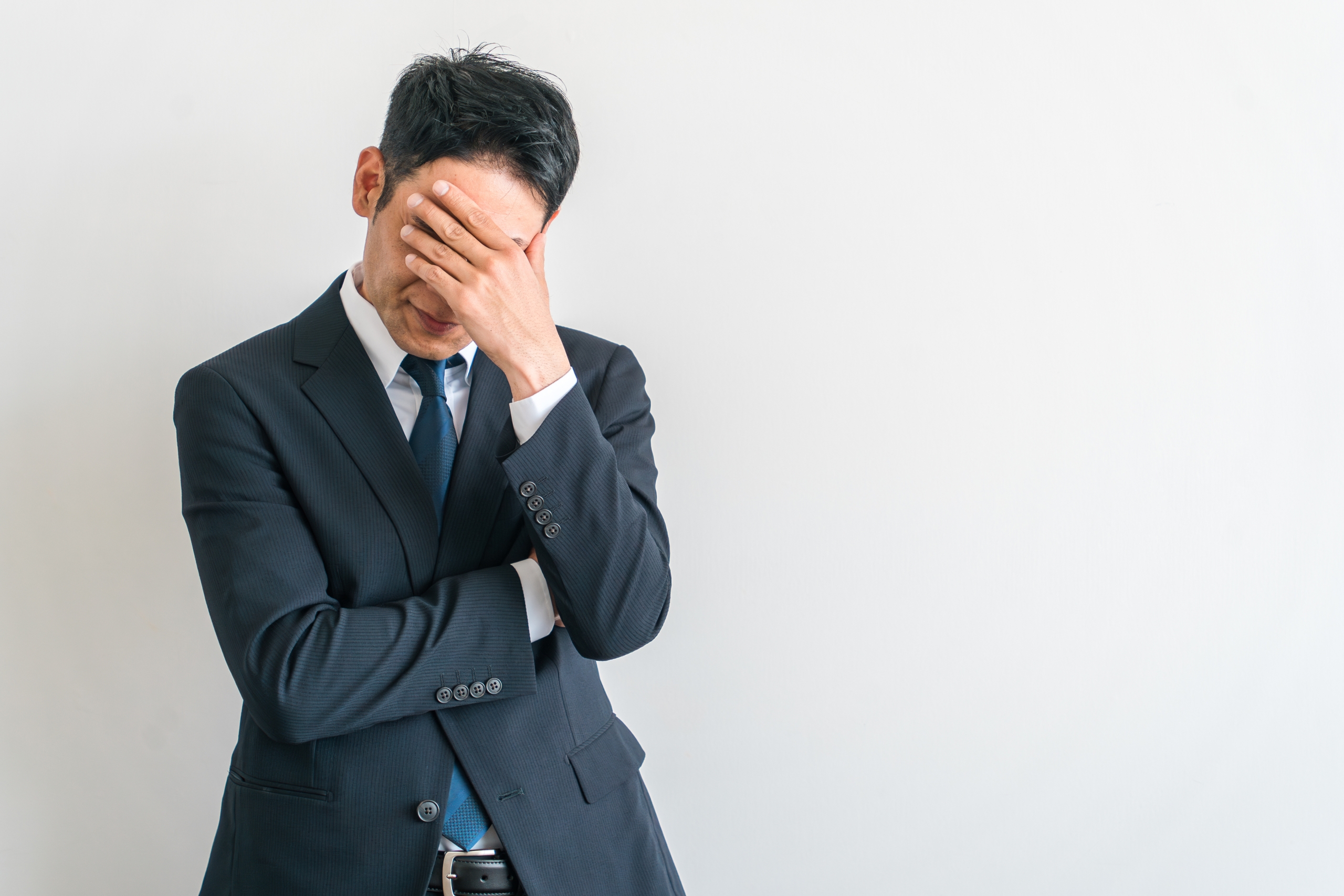 白い背景の前で、顔を手で覆い、ストレスや困惑の表情を見せるスーツ姿のビジネスマン。