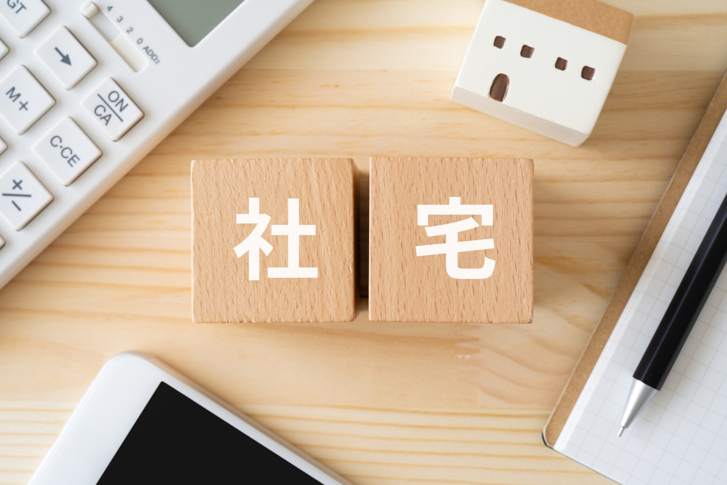木製の積み木に書かれた「社宅」の文字と、電卓やミニチュアの家が置かれたデスク周り