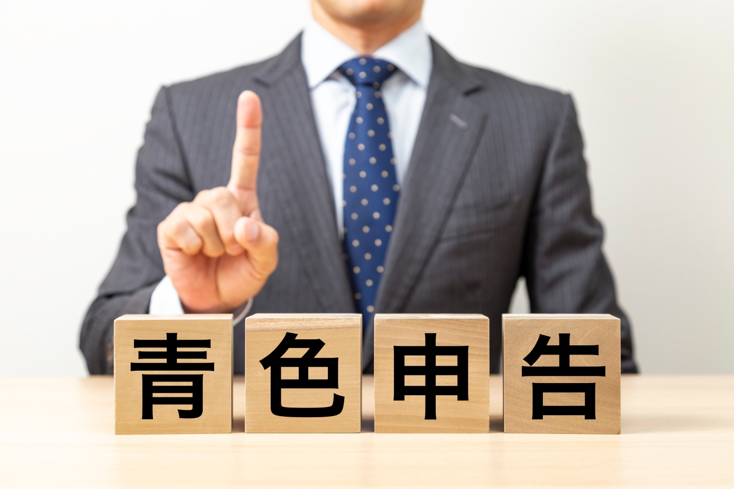 「青色申告」と書かれた木製ブロックを前に指を立てるスーツ姿の人物